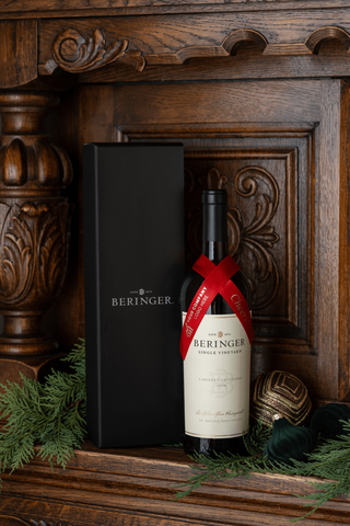 A bottle of Beringer Single Vineyard Cabernet Sauvignon with a red ribbon bow, standing next to its black gift box on a dark wooden mantel decorated with green pine branches and a gold ornament.